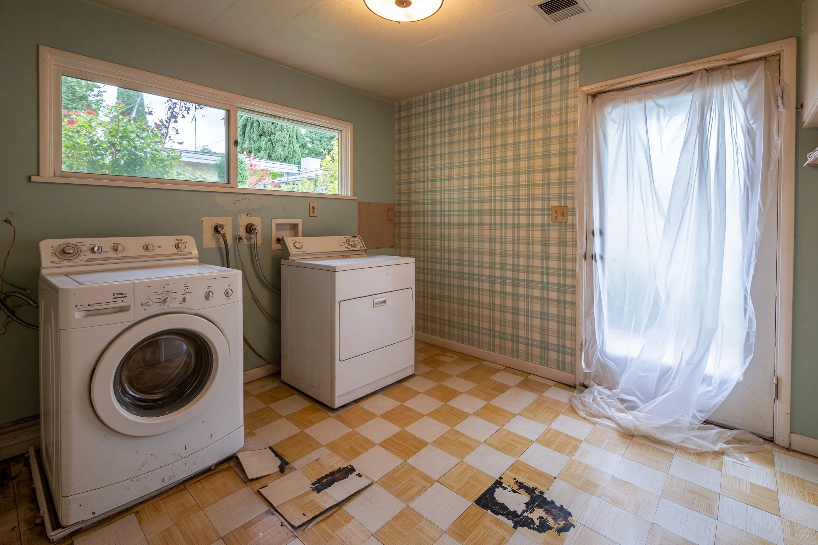 A 1960s laundry room with the washer and dryer still in place and checkered vinyl tile lifted in patches to show black adhesive