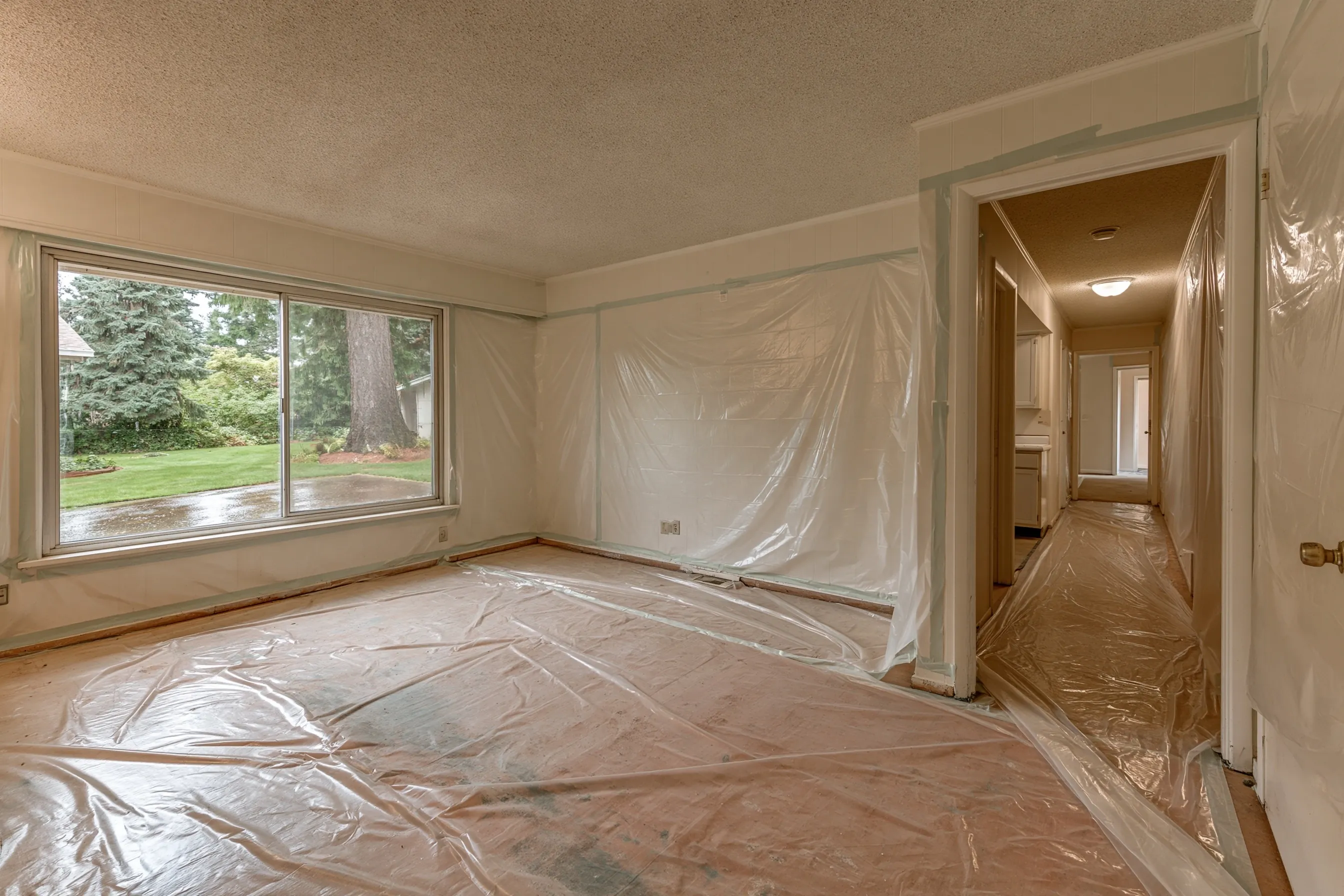 Empty 1970s rambler living room and hall with popcorn ceilings throughout, bare floors, plastic sheeting rolled out on the floor