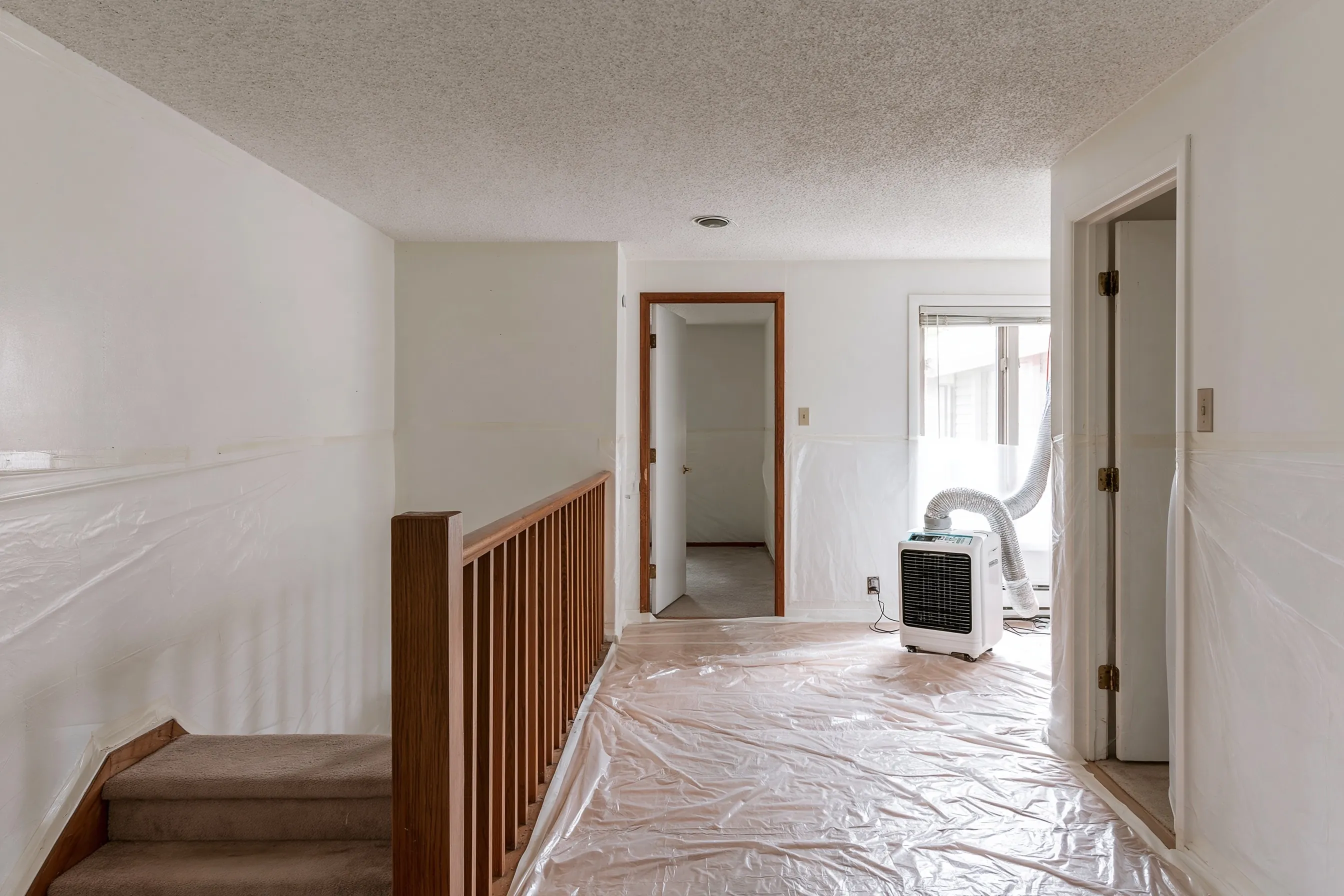 Upper floor of a 1960s split-level with a painted popcorn ceiling, plastic over floor and walls, the half-flight stair in view